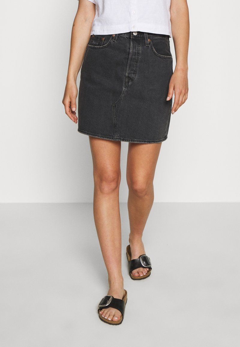 Femme portant une mini-jupe en denim noir, une chemise courte blanche et des sandales noires à enfiler avec de grandes boucles argentées, marchant sur un sol clair.