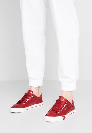 Rode lage sneakers met witte zolen en satijnen veters gedragen met witte joggerbroek op een grijze vloer.