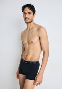 Zwarte katoenen boxershorts met Calvin Klein logo op de tailleband; model staat met een blote torso en kort haar, iets naar de zijkant gewend.