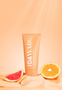 Peachfärgad tub med IDAWARG lätt kroppslotion med vit text, bredvid skivor av apelsin och grapefrukt på mjukt tyg, mot en ljus orange bakgrund.
