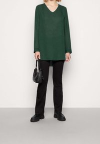 Grön långärmad V-ringad blus med rak passform, kombinerad med svarta åtsittande byxor och kraftiga svarta skor. Håller en liten svart handväska.