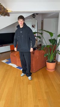 Black hoodie with drawstrings, black pants, and silver shoes. Standing on wooden floor next to a green plant and an orange couch.