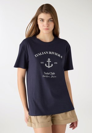 Giovane donna che indossa una maglietta blu navy del Club di Yacht della Riviera Italiana e pantaloncini beige, in piedi davanti a uno sfondo bianco uniforme.