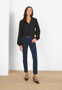 Black blouse with long, puffed sleeves, paired with fitted navy pants and black stiletto heels, on a light wooden floor against a white wall.