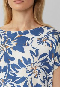 Chemisier floral crème avec un grand imprimé floral bleu et marron. Présente un col bateau et un tissu doux et léger avec des manches courtes.