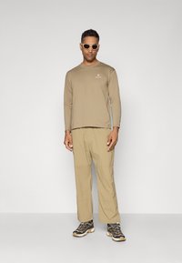 Beige long-sleeve shirt with a white logo, matching beige pants, and black and beige hiking shoes. The shirt has a loose fit, and the sunglasses are gray.