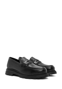 Mocassins en cuir noir avec un profil bas, présentant une texture lisse, une pointe arrondie et une semelle en caoutchouc avec un design de semelle épaisse.
