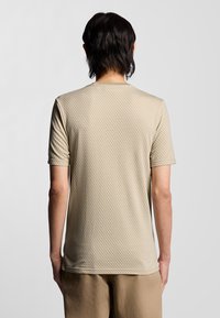 Beige T-Shirt mit kurzen Ärmeln und strukturiertem Muster. Rundhalsausschnitt und lockere Passform, kombiniert mit hellen Hosen.