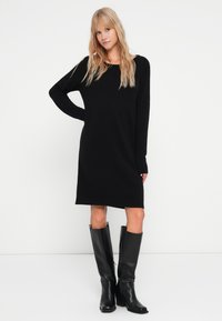 Abito lungo a maniche lunghe nero in maglia con scollatura rotonda, dotato di polsini e bordo a coste. Abbinato a stivali neri in pelle con tacco alti fino al ginocchio.