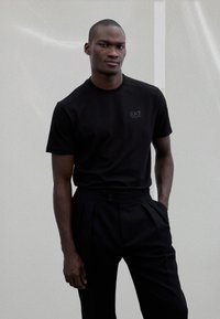 Juoda trumpomis rankovėmis marškinėliai su subtiliu EA7 logotipu, derinami su juodomis, aukštu liemeniu, klostuotomis kelnėmis, puoštomis dekoratyvinėmis sagomis.