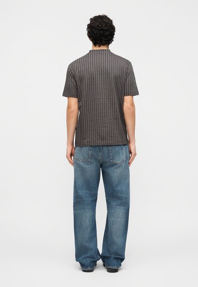 Άνδρας με σγουρά μαλλιά, που φοράει κοντομάνικο πουκάμισο με σχέδια και άνετο μπλε τζιν, στέκεται αντικρίζοντας ένα απλό λευκό φόντο.