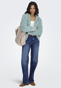 Hellblaue, flauschige Jacke mit Reißverschluss über einem weißen Oberteil, kombiniert mit blauen, ausgestellten Jeans und braunen Slippern. Inklusive großer beiger Tragetasche.