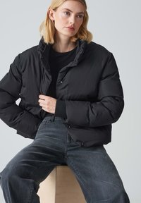 Giubbotto imbottito nero con colletto alto, bottoni a scatto e design cropped, abbinato a jeans in denim scuro, seduto su una superficie di legno.