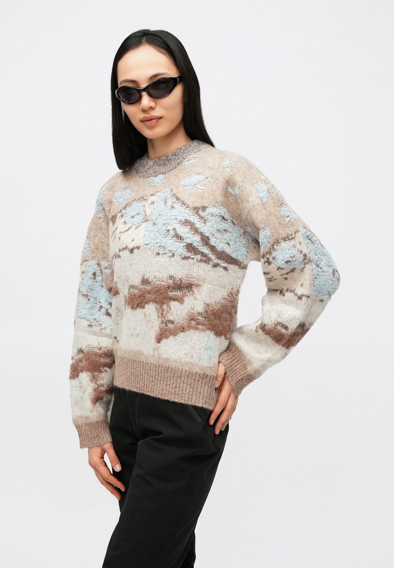 Maglione lavorato a maglia beige con motivi astratti celesti e marroni. Presenta una scollatura rotonda e polsini a coste. Indossato con pantaloni neri.