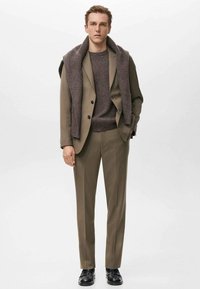 Brązowy garnitur z prążkowanym brązowym swetrem pod spodem, dopasowany do tego szalik zawieszony na ramionach. Czarny buty uzupełniają stylizację.