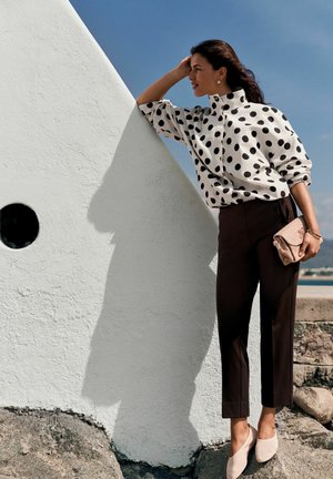 Woman in black pants and white polka-dot blouse poses by white wall, holding beige clutch, against clear blue sky background.