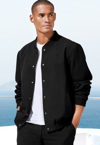 Black bomber jacket made of soft fabric with snap buttons, ribbed cuffs, and waistband, worn over a white t-shirt, set against a coastal backdrop.