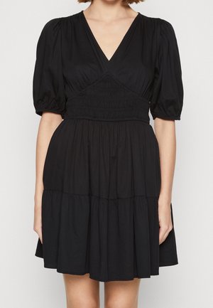 Kobieta ubrana w krótką czarną sukienkę z bufiastymi rękawami, dekoltem w serek, marszczoną talią i wielowarstwową spódnicą.