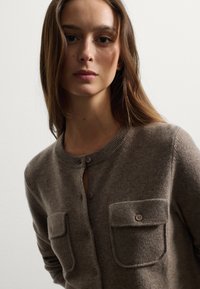 Cárdigan de punto de color marrón claro con escote redondo, dos bolsillos en el pecho y cierre de botones. Textura suave con bordes acanalados.