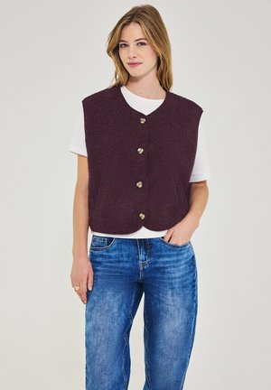 Kvinde iført en hvid kortærmet skjorte, lilla tekstureret vest med knapper og blå jeans, stående med den ene hånd i lommen mod en hvid baggrund.