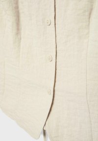 Gros plan sur le devant d'une chemise en lin beige montrant la texture du tissu et trois petits boutons ronds attachés le long de la patte de boutonnage.