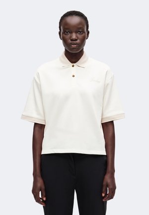 Femme aux cheveux courts portant un polo blanc à manches courtes avec une bordure beige et un pantalon noir, debout devant un fond clair uni.