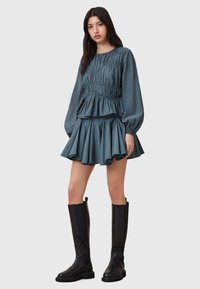 Top azzurro con maniche lunghe a sbuffo abbinato a una gonna mini plissettata. Il modello indossa stivali neri al ginocchio. Design semplice e strutturato.