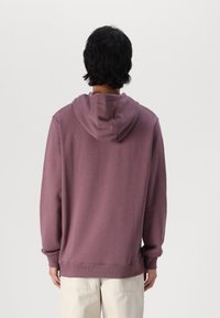 Sudadera de algodón púrpura con capucha ajustable, puños y dobladillo acanalados, y un corte holgado, llevada sobre pantalones de color crema.
