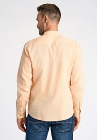 Camicia lunga a maniche con bottoni color pesca chiaro, in tessuto testurizzato e orlo arrotondato, indossata con jeans blu, che mostrano una vestibilità rilassata.