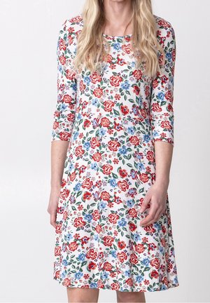 Kobieta ubrana w białą sukienkę w czerwono-niebiesko-zielony kwiatowy wzór, z rękawami do połowy przedramienia i okrągłym dekoltem.