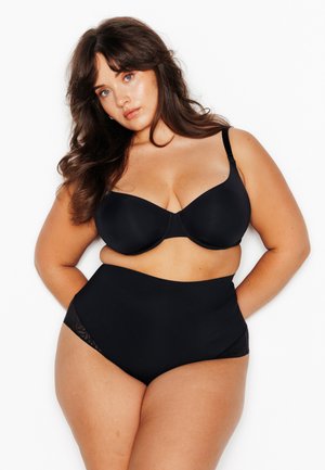 Femme aux longs cheveux bruns portant un soutien-gorge noir et une culotte noire taille haute sur un fond blanc uni.