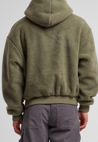 Personne portant une veste polaire verte à capuche avec poignets et ourlet côtelés, associée à un pantalon en velours côtelé gris, vue de dos.