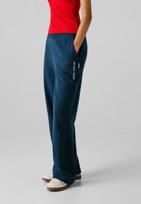 Osoba ubrana w granatowe spodnie "Red Bull Racing" z kieszeniami, czerwoną bluzkę i białe trampki na jasnoszarym tle.