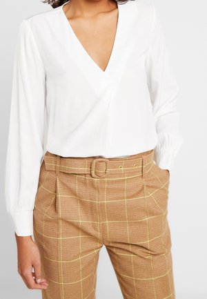 Vrouw draagt een witte blouse met lange mouwen en V-hals, ingestopt in een bruine geruite hoge taille broek met bijpassende riem, handen in de zakken.