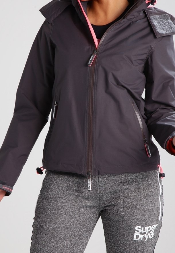 Osoba nosící tmavě šedou bundu na zip s růžovými detaily a šedé tepláky SuperDry s bílým logem na stehně.