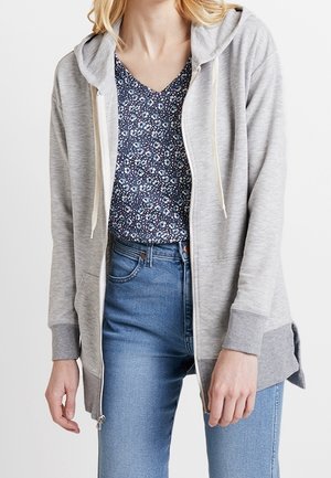 Felpa grigia con zip e cappuccio regolabile, dotata di maniche lunghe e spacchi laterali, indossata su una maglietta a scollo a V con motivo floreale scuro e jeans blu.