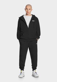 Czarna bluza z kapturem na zamek, z sznurkiem i kieszeniami, połączona z czarnymi dresami. Model nosi białą koszulę i białe sneakersy z kolorowymi akcentami.