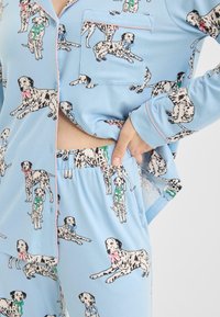 Blaues Pyjama-Set mit einem Muster aus Dalmatinern mit Schleifen, gefertigt aus glattem Stoff, bestehend aus einem Oberteil mit Knopfverschluss und Tasche sowie einem Hosenbund mit elastischem Bund.