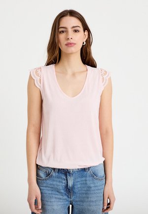 Jonge vrouw draagt een lichtroze mouwloze top met kanten rand en blauwe spijkerbroek, staand tegen een effen witte achtergrond.