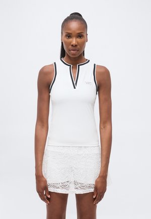 Femeie purtând un top alb fără mâneci cu margine neagră și o fustă albă din dantelă, stând cu fața spre înainte pe un fundal simplu.