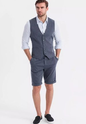 Veste en tissu texturé marine avec fermeture à boutons, associée à un short à ourlet roulé. Chemise blanche à rayures fines en dessous, complétée par des chaussures noires à enfiler.