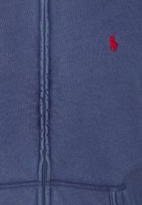 Sudadera con cremallera en azul marino hecha de tela suave, con cremallera central, dobladillo acanalado y un pequeño logo bordado en rojo en el pecho.