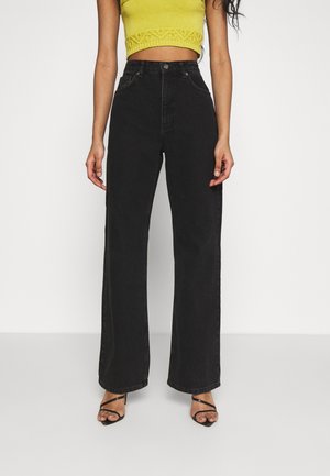 Femme portant un jean noir taille haute à jambes larges, un top court sans manches jaune et des talons ouverts noirs à lanières, debout devant un fond blanc.