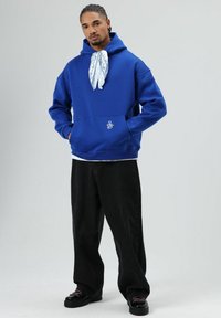 Blauwe hoodie met een voorzak, voorzien van een wit logo en een sjaal met patroon om de hals geknoopt. Gecombineerd met zwarte broek met wijde pijpen.