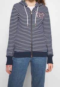Felpa con zip a righe blu navy e bianche, dotata di cappuccio, con lacci bianchi, polsini a coste e dettaglio del logo sul davanti. Vestibilità casual.
