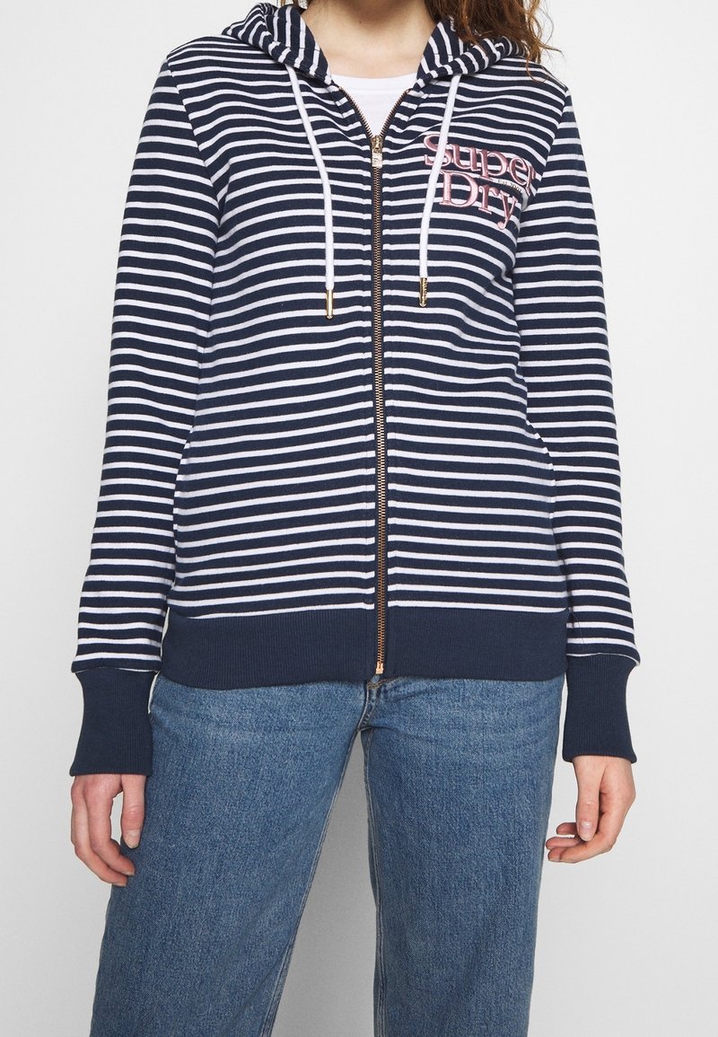 Felpa con zip a righe blu navy e bianche, dotata di cappuccio, con lacci bianchi, polsini a coste e dettaglio del logo sul davanti. Vestibilità casual.