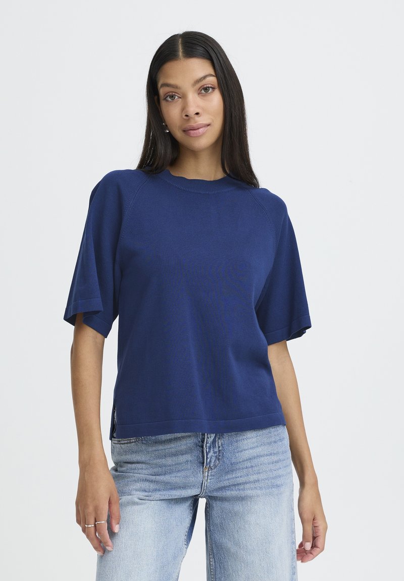 Žena s dlouhými tmavými vlasy, oblečená v volné navy modré blůze s krátkým rukávem a světle modrých džínách, na jednobarevném bílém pozadí.