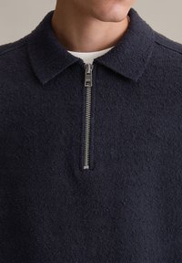 Granatowy, teksturowany sweter z kołnierzykiem i zamkiem błyskawicznym na ćwierć długości. Materiał wydaje się miękki i puszysty, z subtelnym, nierównym wzorem na powierzchni.