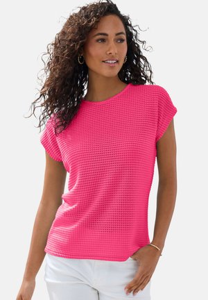 Woman with curly hair wearing a bright pink textured short-sleeve top and white pants, posing with one hand in pocket.