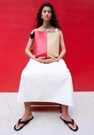 Mujer con cabello largo y rizado sentada en una silla junto a una pared roja, vestida con vestido blanco y chanclas negras, sosteniendo un bolso grande rojo y beige.